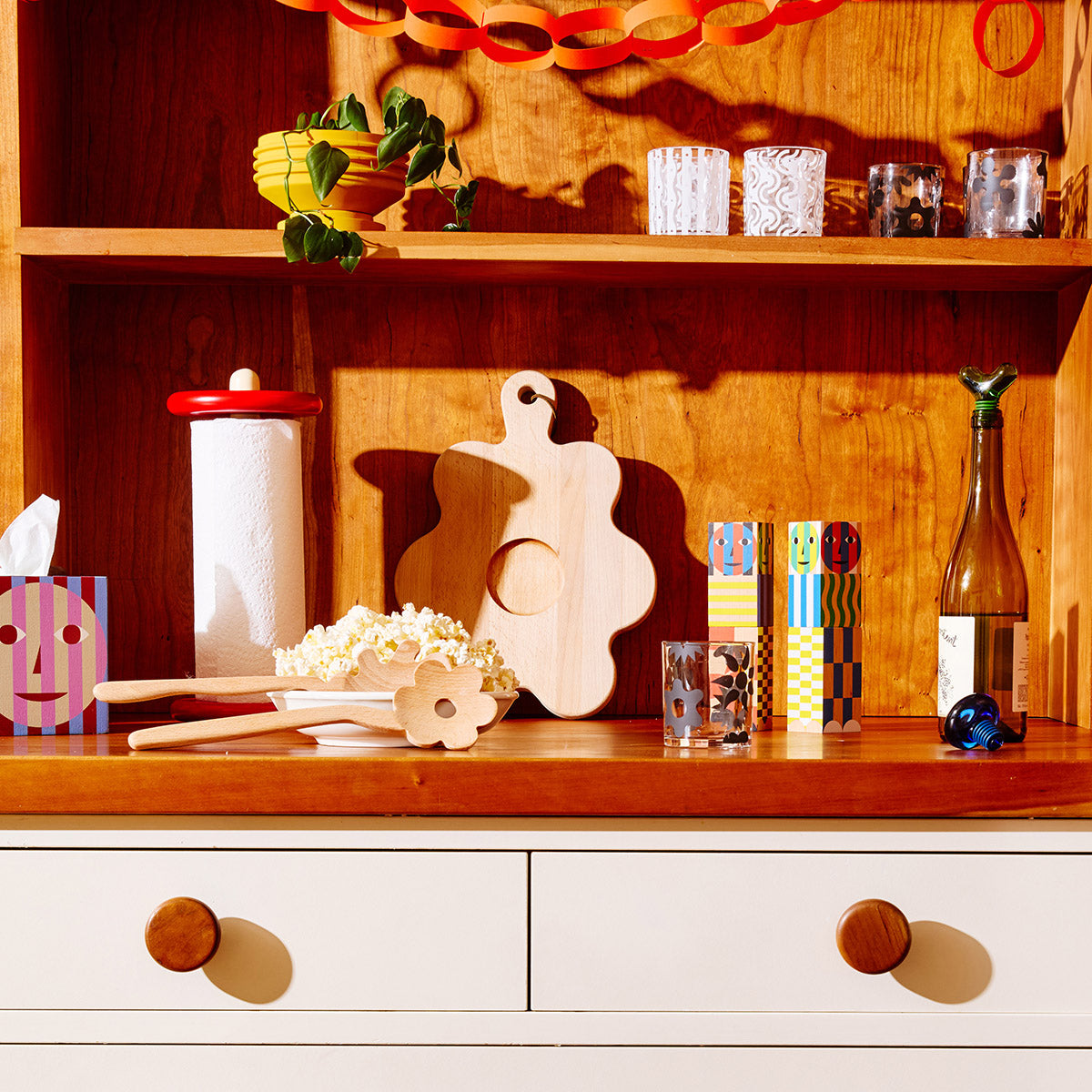 Colorful kitchen accessories sit atop a wooden countertop. Items include a red paper towel holder, a flower-shaped cutting board, and salt and pepper grinders with colorful patterns.