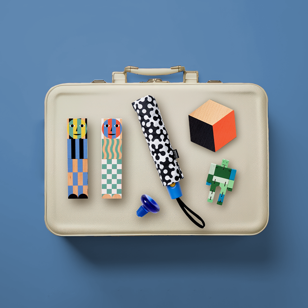 Small items like an umbrella, kitchen accessories, and toys lay on top of a cream suitcase with a blue background.