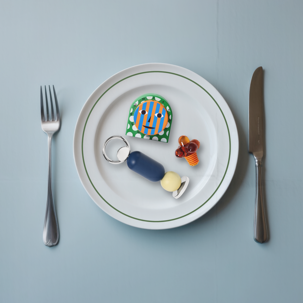 Plate with colorful toy-like items, fork, knife, and a glass bottle stopper on a light blue background.
